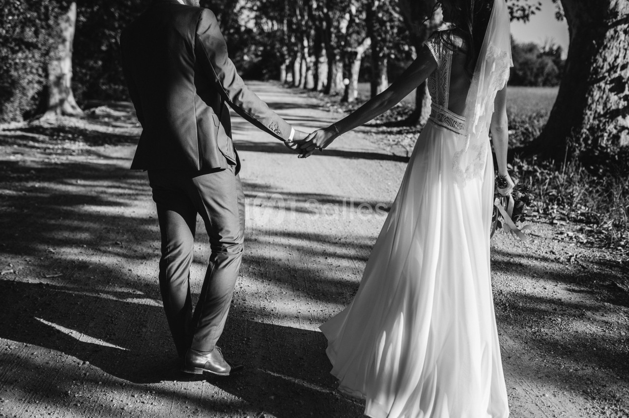 Un couple en tenue de mariage marche main dans la main sur un chemin bordé d'arbres.