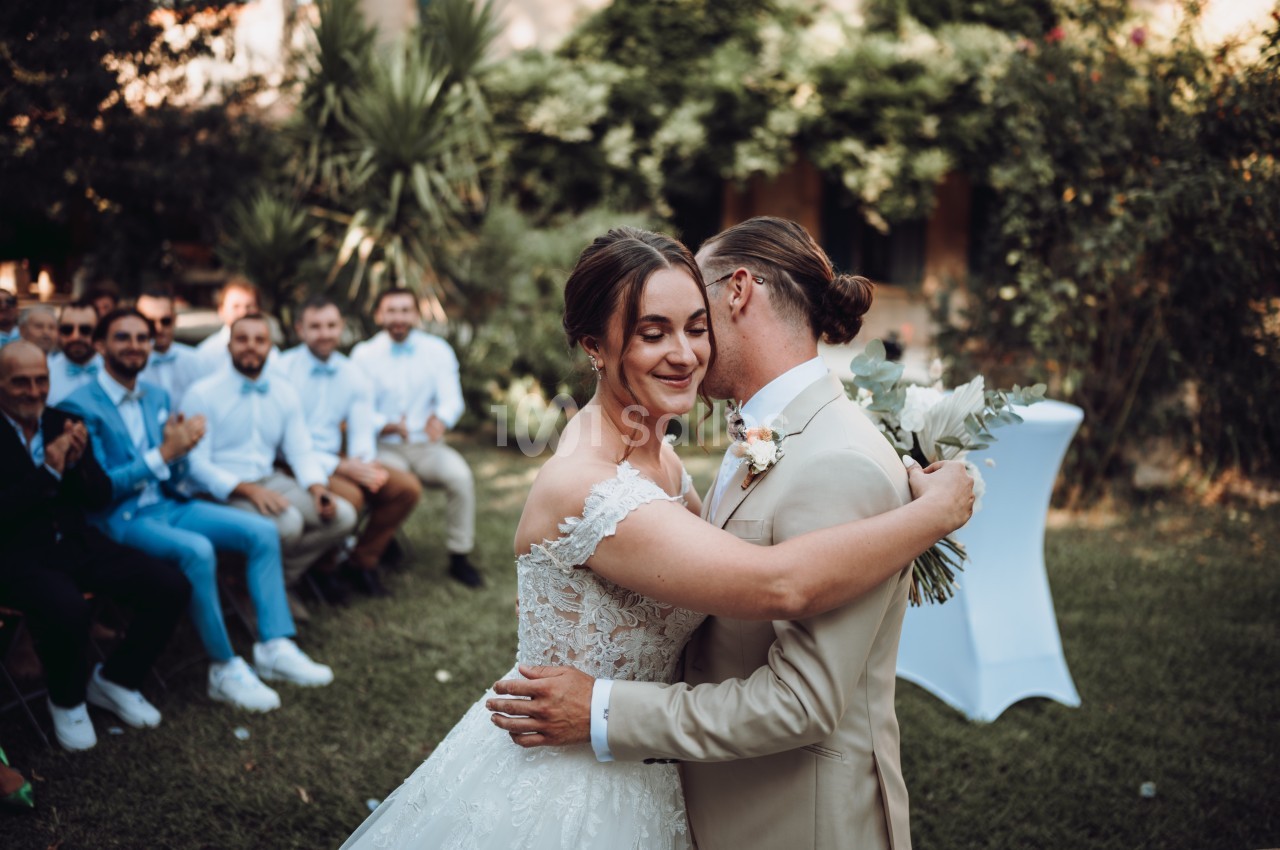 Un couple en tenue de mariage s'enlace dans un jardin, entouré d'invités assis.