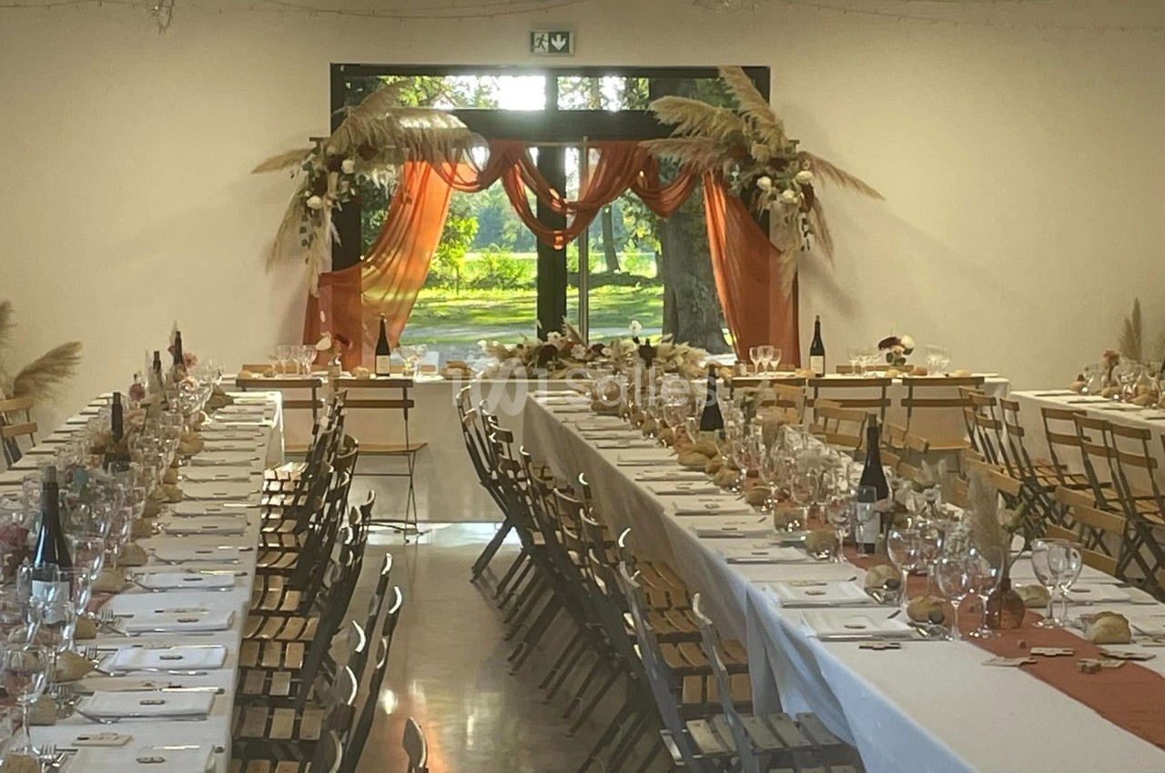 Salle décorée pour un événement avec longues tables dressées, nappes blanches et arrangements floraux près d'une baie vitrée.
