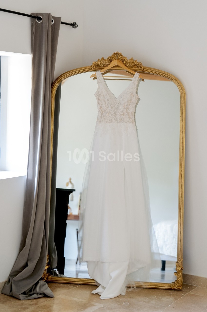 Robe de mariée blanche suspendue à un cintre devant un grand miroir doré dans une pièce lumineuse.