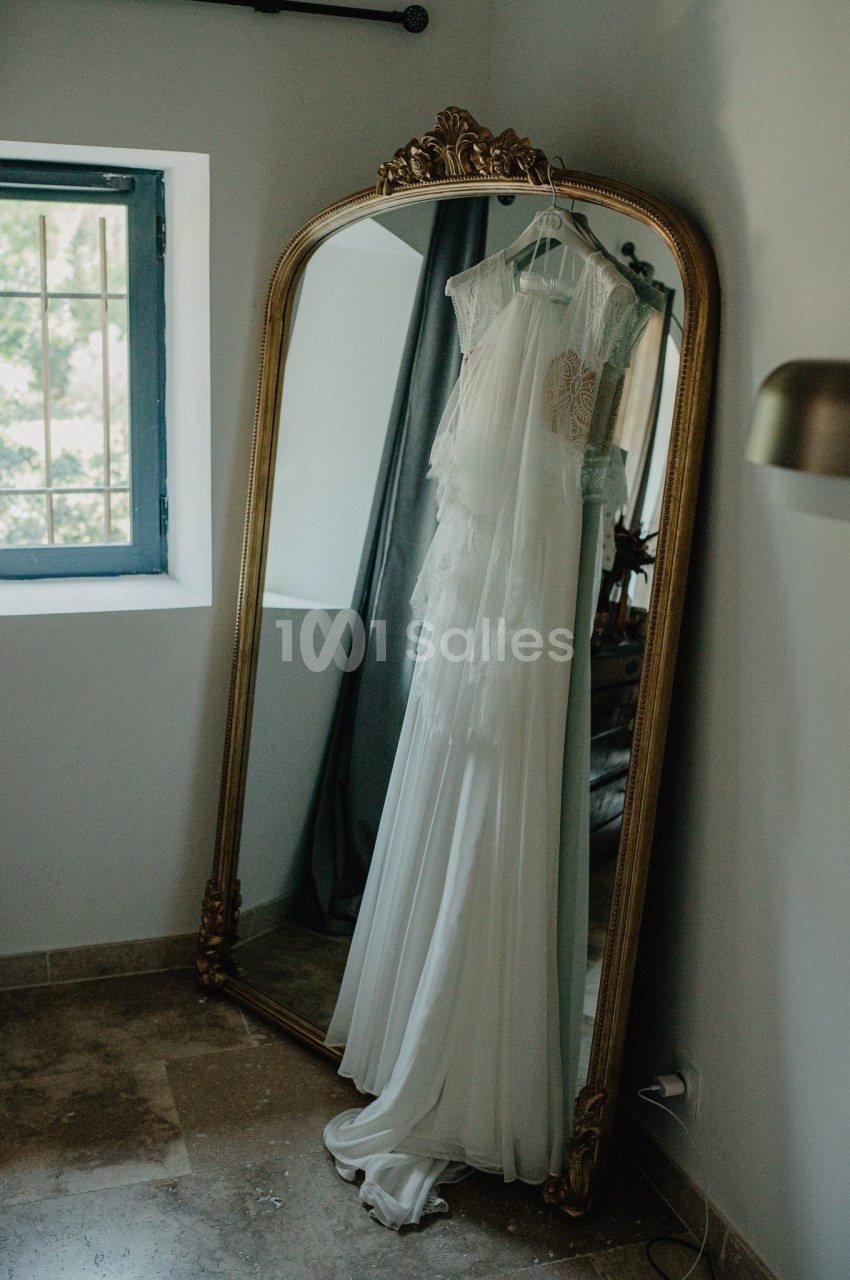 Robe de mariée blanche suspendue à un miroir doré dans une pièce lumineuse avec fenêtre et rideaux.