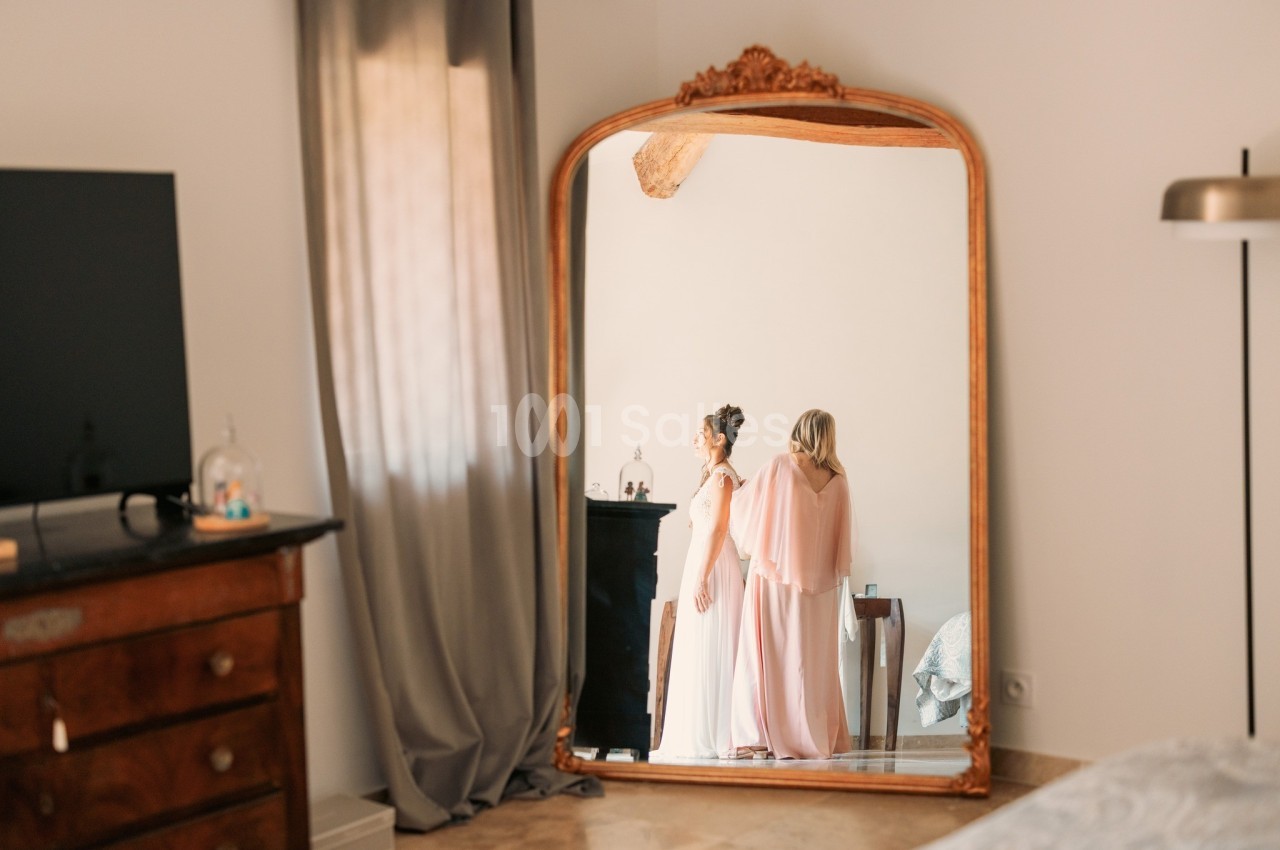 Deux femmes en robes élégantes se tiennent devant un grand miroir encadré de bois dans une chambre lumineuse.