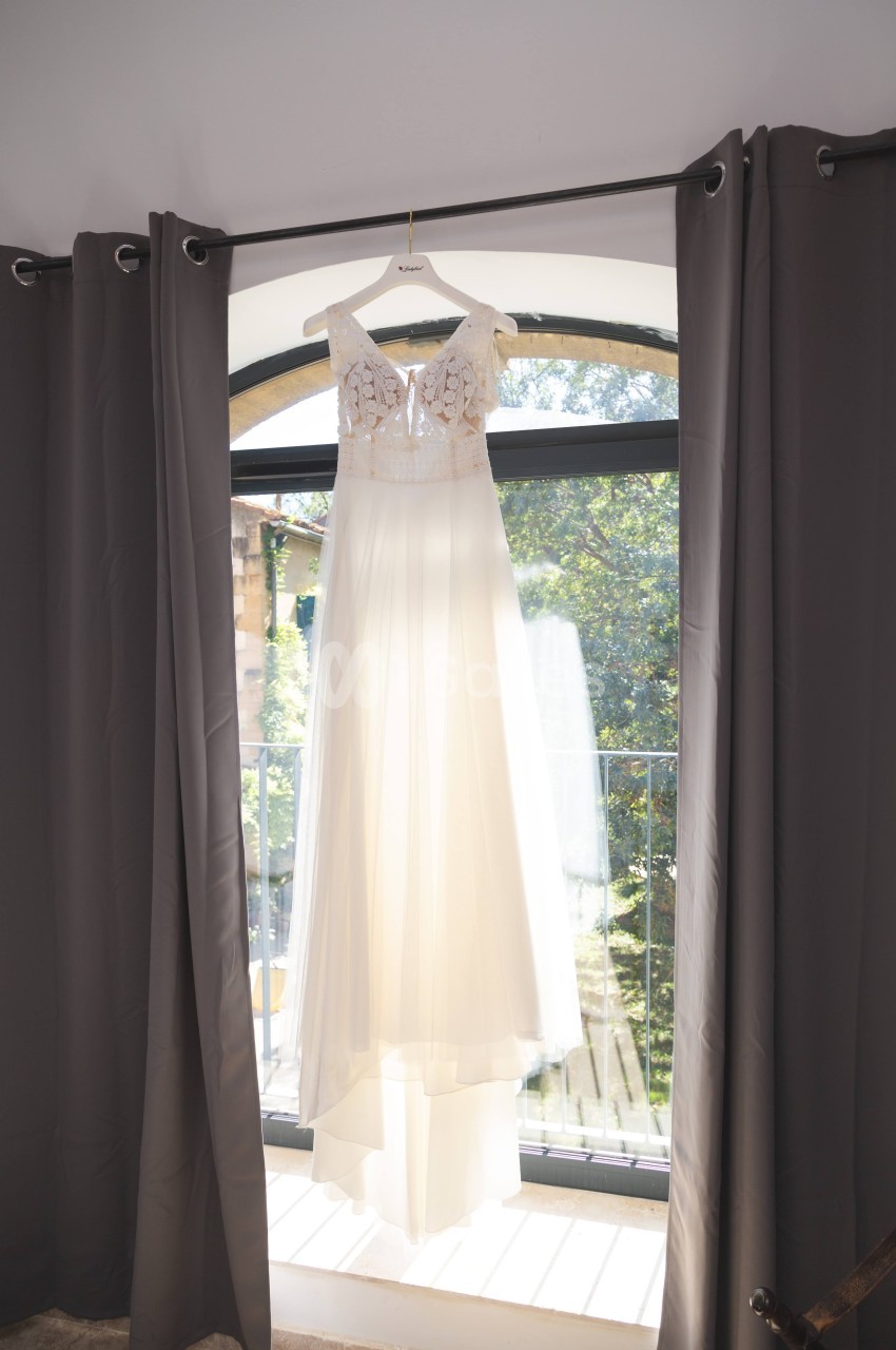 Robe de mariée blanche suspendue à un cintre devant une fenêtre encadrée de rideaux gris.
