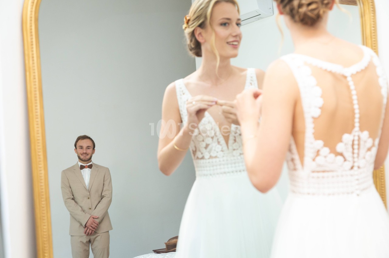 Une femme en robe de mariée s'observe dans un miroir, un homme en costume beige se tient en arrière-plan.