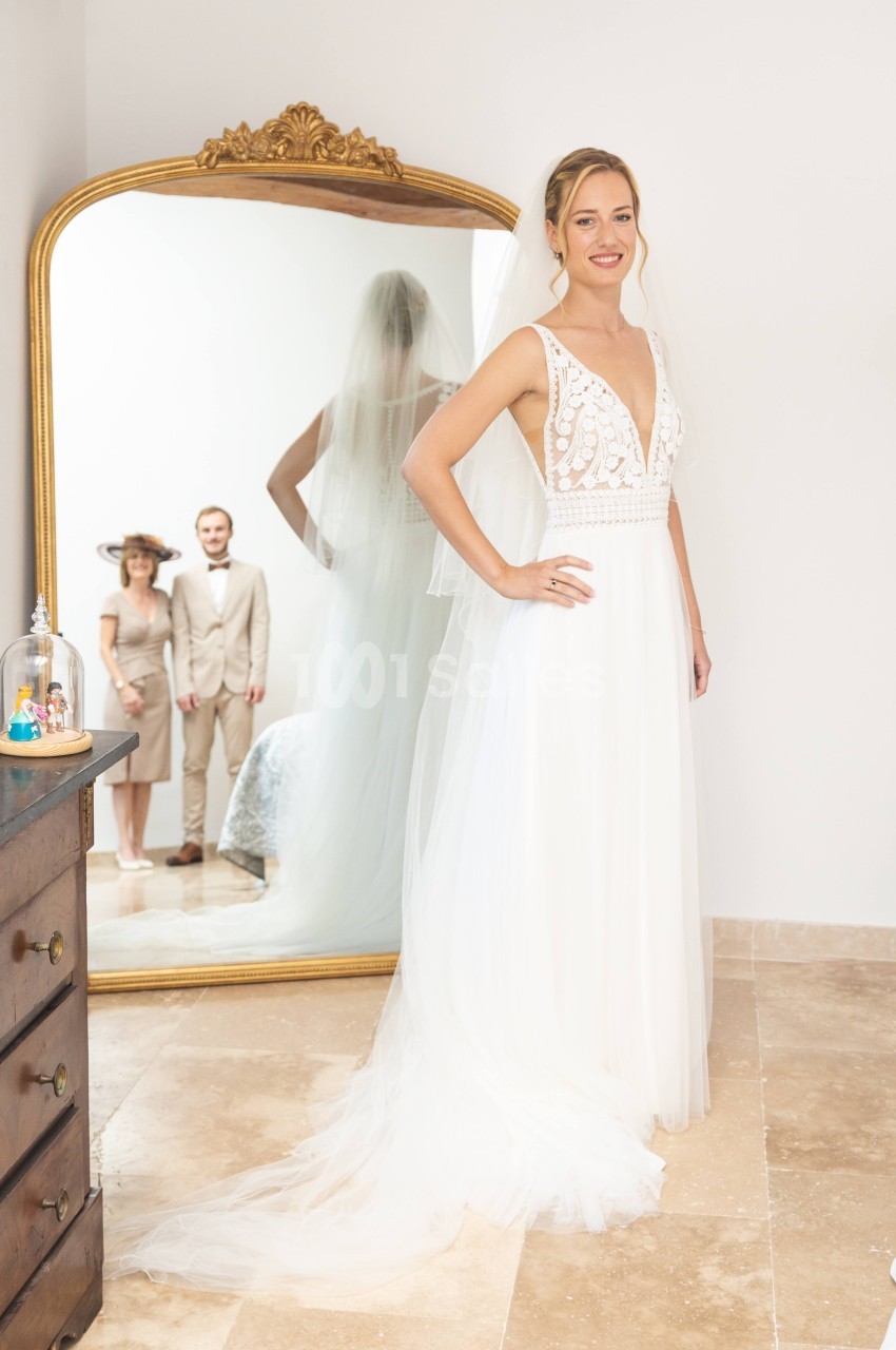 Une mariée en robe blanche se tient devant un miroir, reflétant un couple en arrière-plan dans une pièce lumineuse.