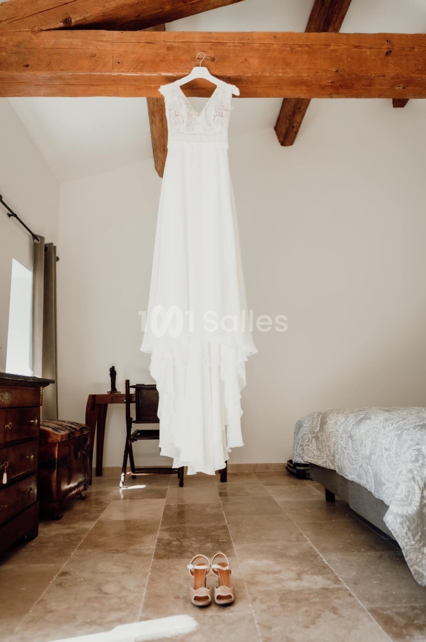 Robe de mariée blanche suspendue à une poutre en bois dans une chambre lumineuse, avec des chaussures posées au sol.