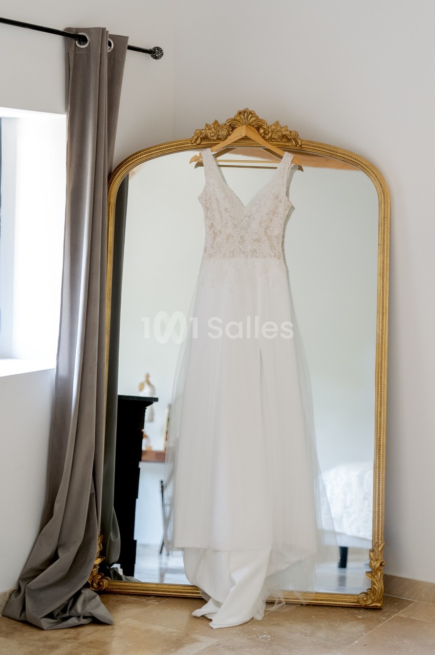 Robe de mariée blanche suspendue à un miroir doré dans une pièce lumineuse avec des rideaux gris.