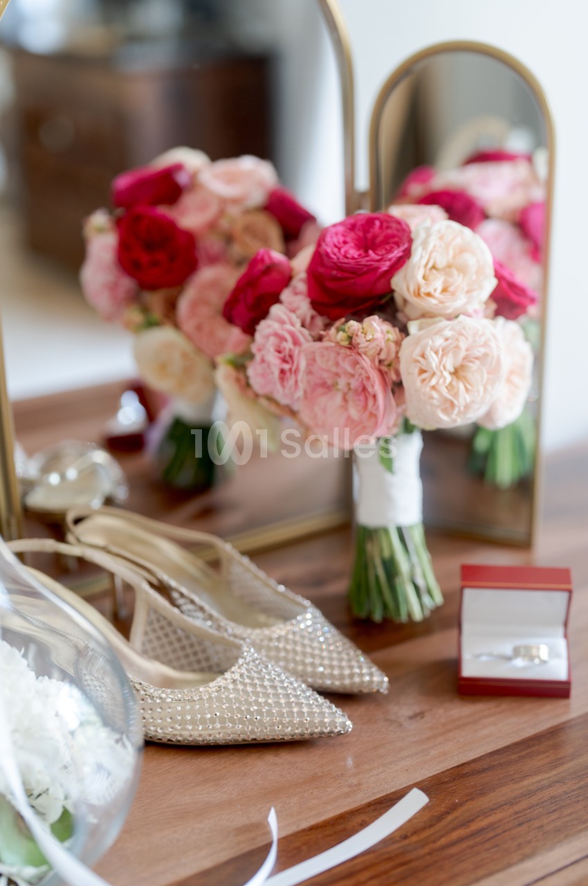 Bouquet de fleurs roses et rouges, escarpins perlés et boîte à alliances posés sur une table devant un miroir.