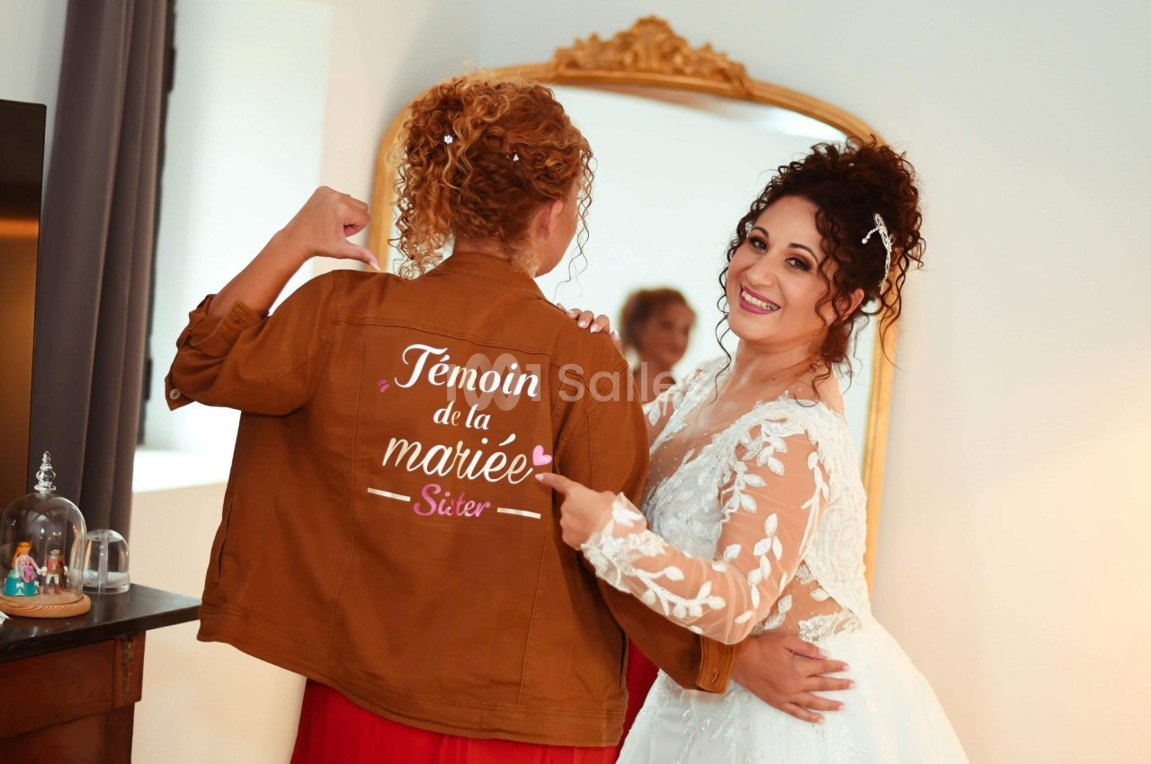 Une mariée souriante pointe la veste de sa témoin, où est inscrit ’Témoin de la mariée, sister’.