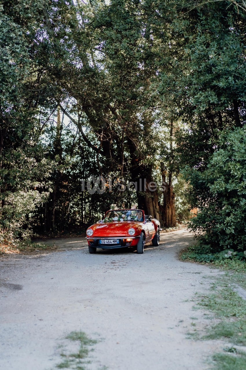 Voiture décapotable rouge garée sur un chemin de terre entouré d'arbres et de végétation dense.