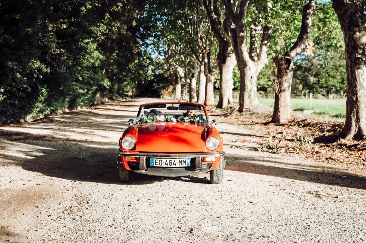 Voiture décapotable rouge circulant sur une route de campagne bordée d'arbres sous un ciel ensoleillé.
