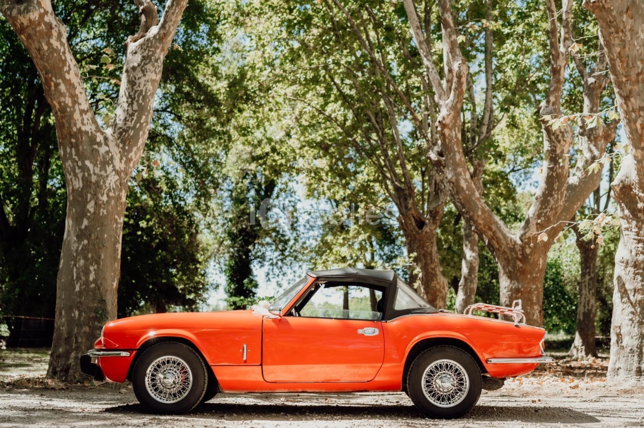 Voiture ancienne rouge décapotable garée sous des arbres dans un environnement ensoleillé.