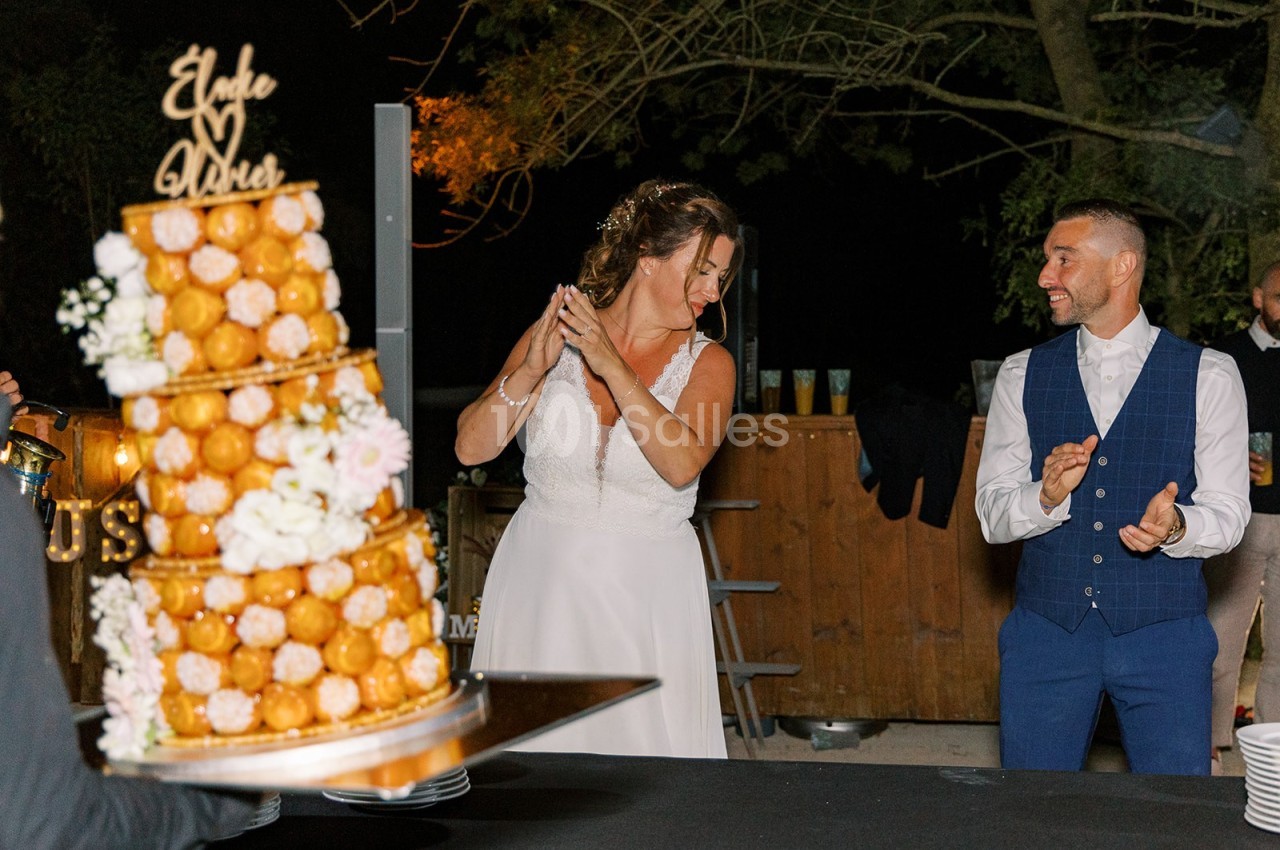 Une mariée et un marié souriants près d'une pièce montée décorée de fleurs, lors d'une réception en extérieur.