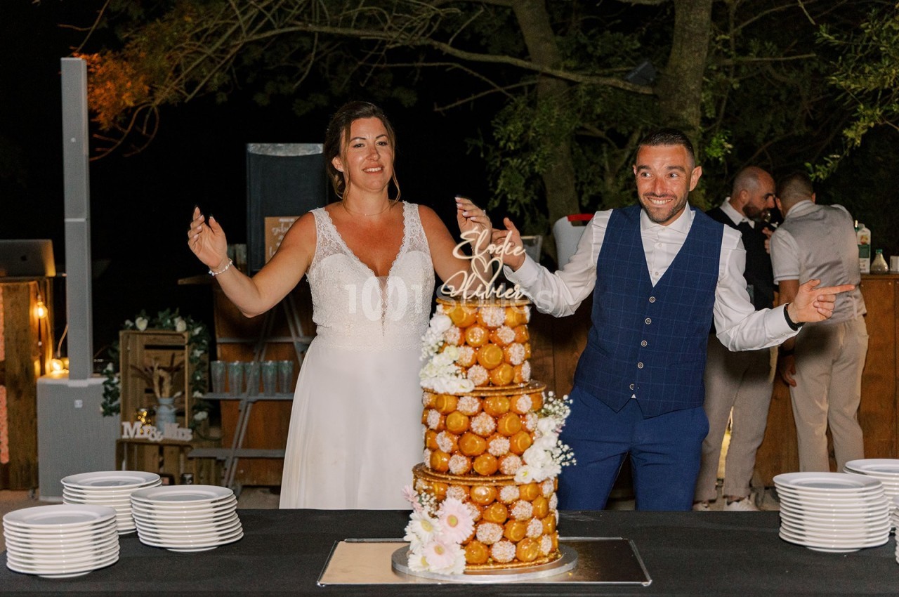Un couple souriant pose devant une pièce montée de choux décorée de fleurs lors d'une réception en extérieur.