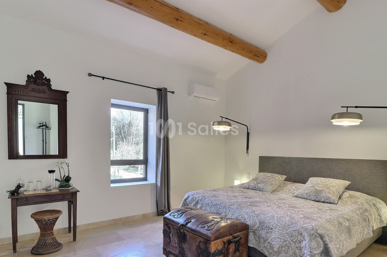 Chambre lumineuse avec lit double, poutres apparentes, miroir ancien, fenêtre donnant sur l'extérieur et éclairage moderne.