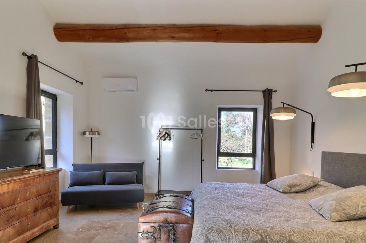 Chambre lumineuse avec lit double, canapé, commode en bois, télévision, et fenêtres donnant sur un paysage extérieur.