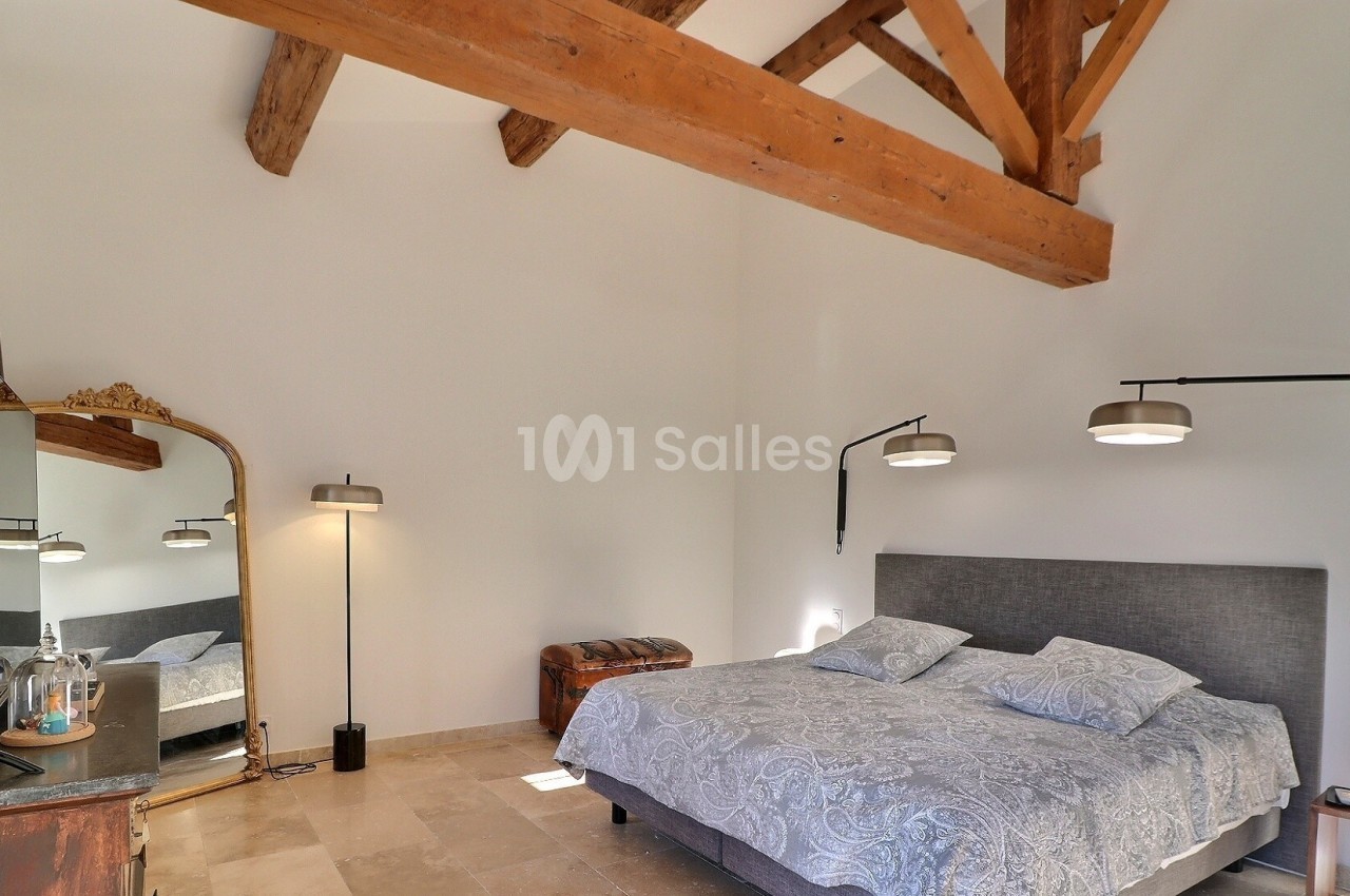 Chambre lumineuse avec lit double, poutres apparentes, miroir sur pied, lampes modernes et sol en pierre beige.