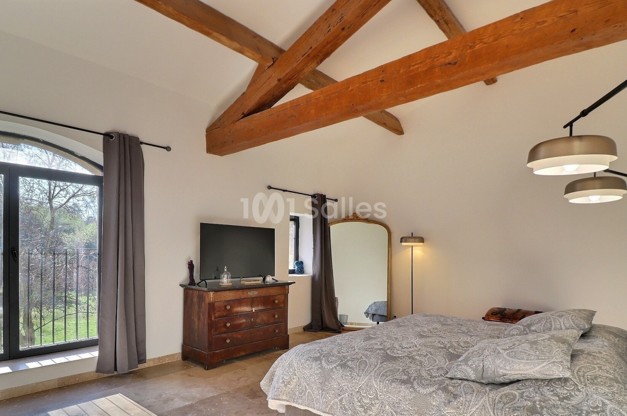 Chambre lumineuse avec poutres apparentes, lit double, commode, miroir, lampadaire et grande fenêtre donnant sur un jardin.