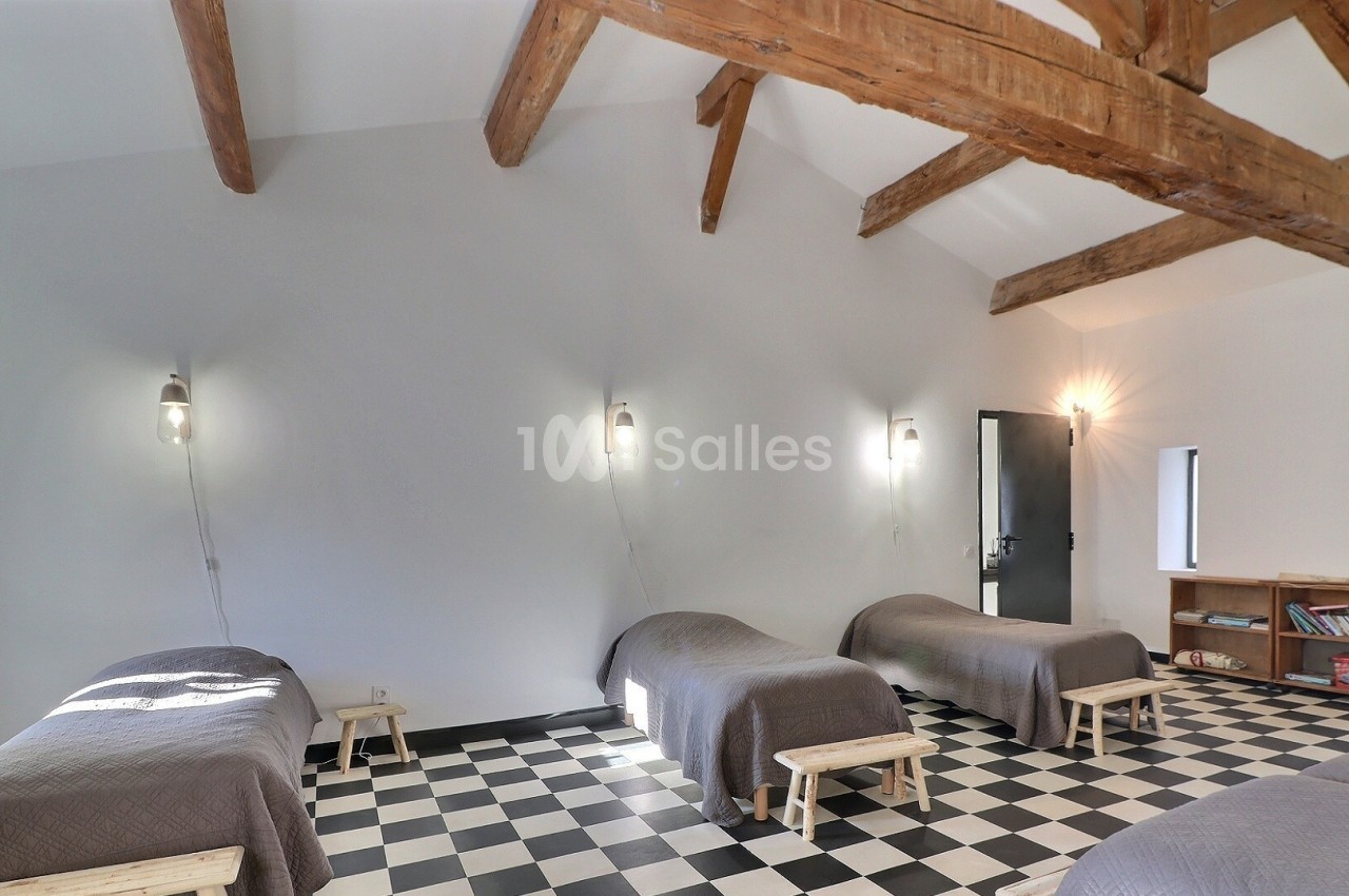 Quatre lits simples alignés dans une pièce lumineuse avec plafond en poutres apparentes et sol en damier noir et blanc.