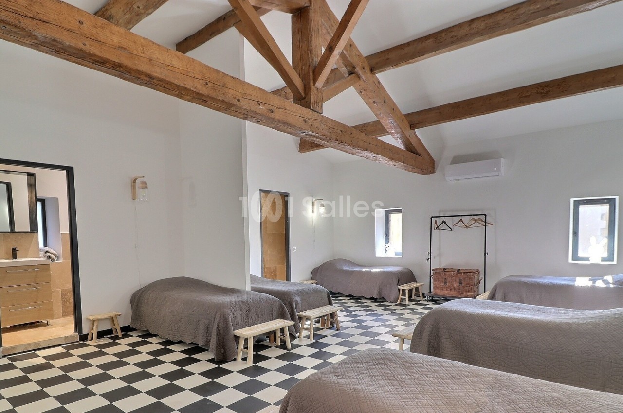 Dortoir lumineux avec poutres apparentes, lits simples alignés, sol en damier et accès à une salle de bain.