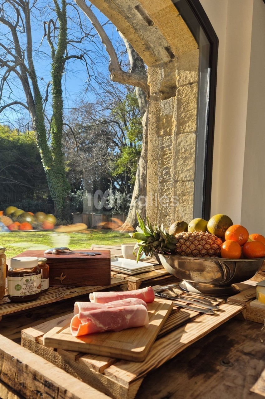 Petit-déjeuner sur une table en bois avec charcuterie, fruits, confitures et vue sur un jardin ensoleillé par une grande…