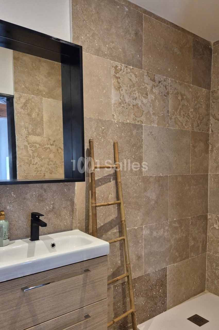 Salle de bain avec lavabo, miroir noir, échelle en bois décorative et murs en carrelage beige texturé.