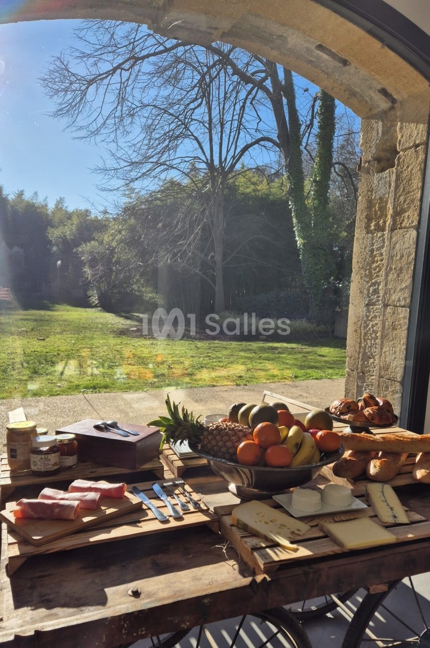 Table en bois avec charcuterie, fromages, fruits et pain, placée devant une baie vitrée donnant sur un jardin ensoleillé.