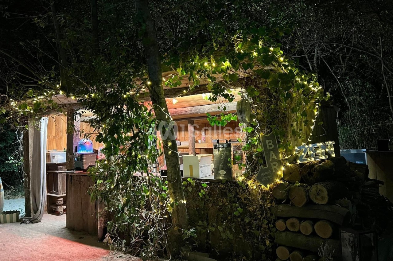 Un bar extérieur en bois décoré de guirlandes lumineuses et entouré de végétation, éclairé de nuit.