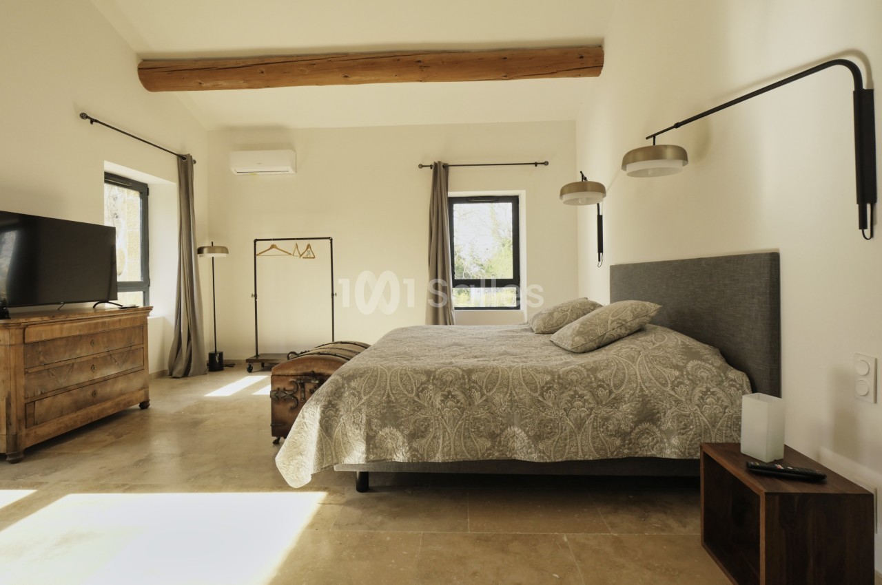 Chambre lumineuse avec un lit double, mobilier en bois, murs clairs, poutre apparente et grandes fenêtres.