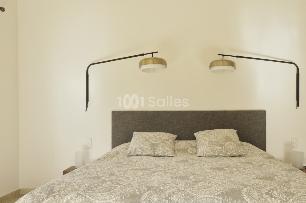 Lit double avec tête de lit grise, draps à motifs paisley et deux lampes murales orientables sur un mur beige.