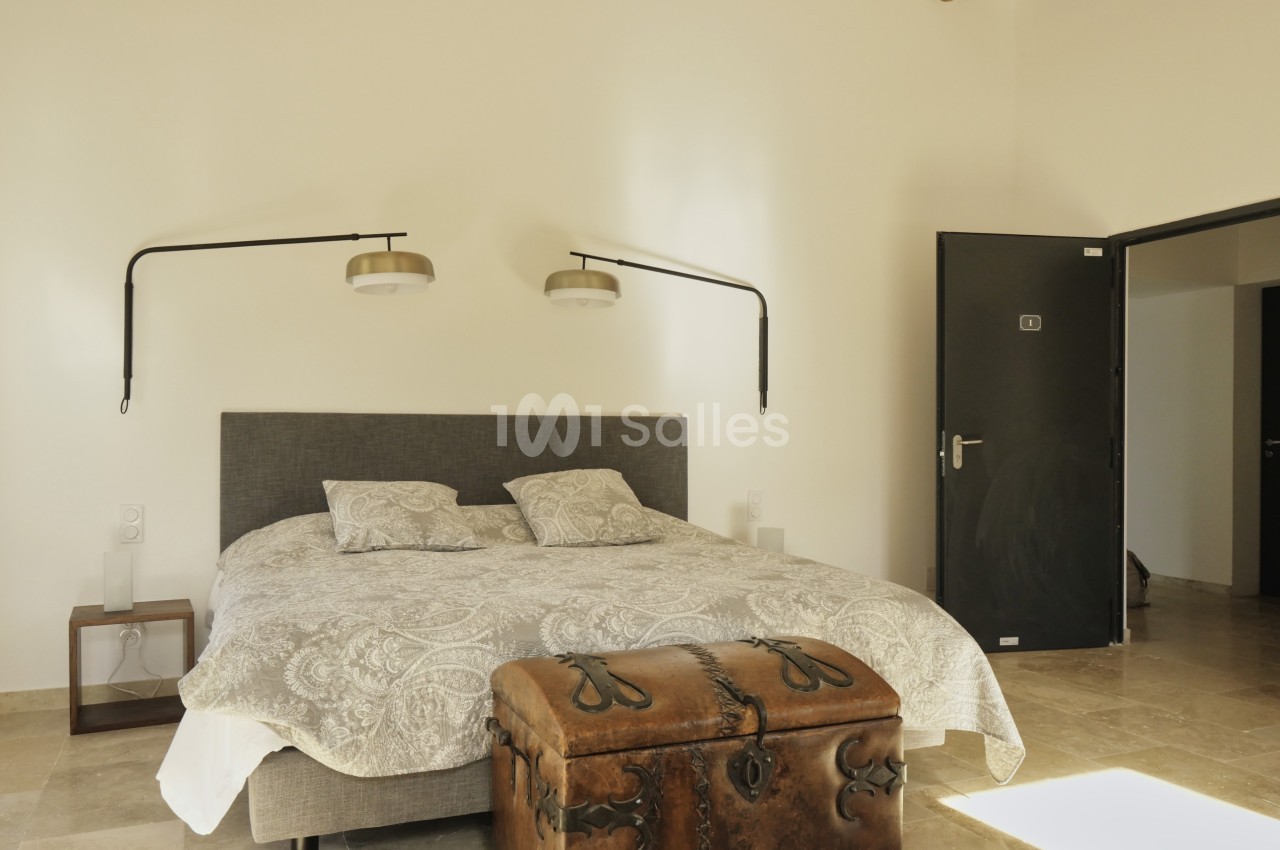 Chambre lumineuse avec un lit double, deux appliques murales, un coffre en bois et un sol en pierre beige.