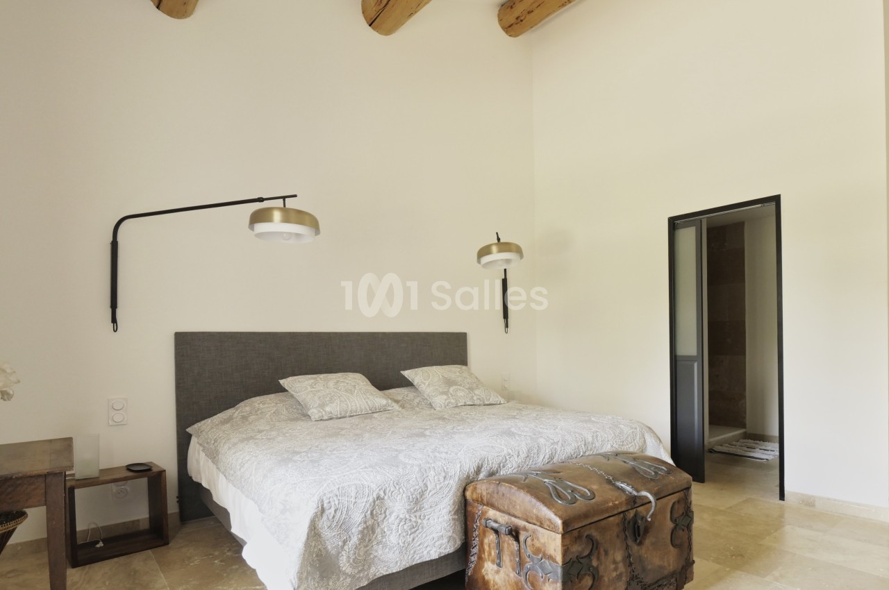 Chambre lumineuse avec lit double, poutres apparentes, murs blancs, sol en pierre et coffre en bois au pied du lit.