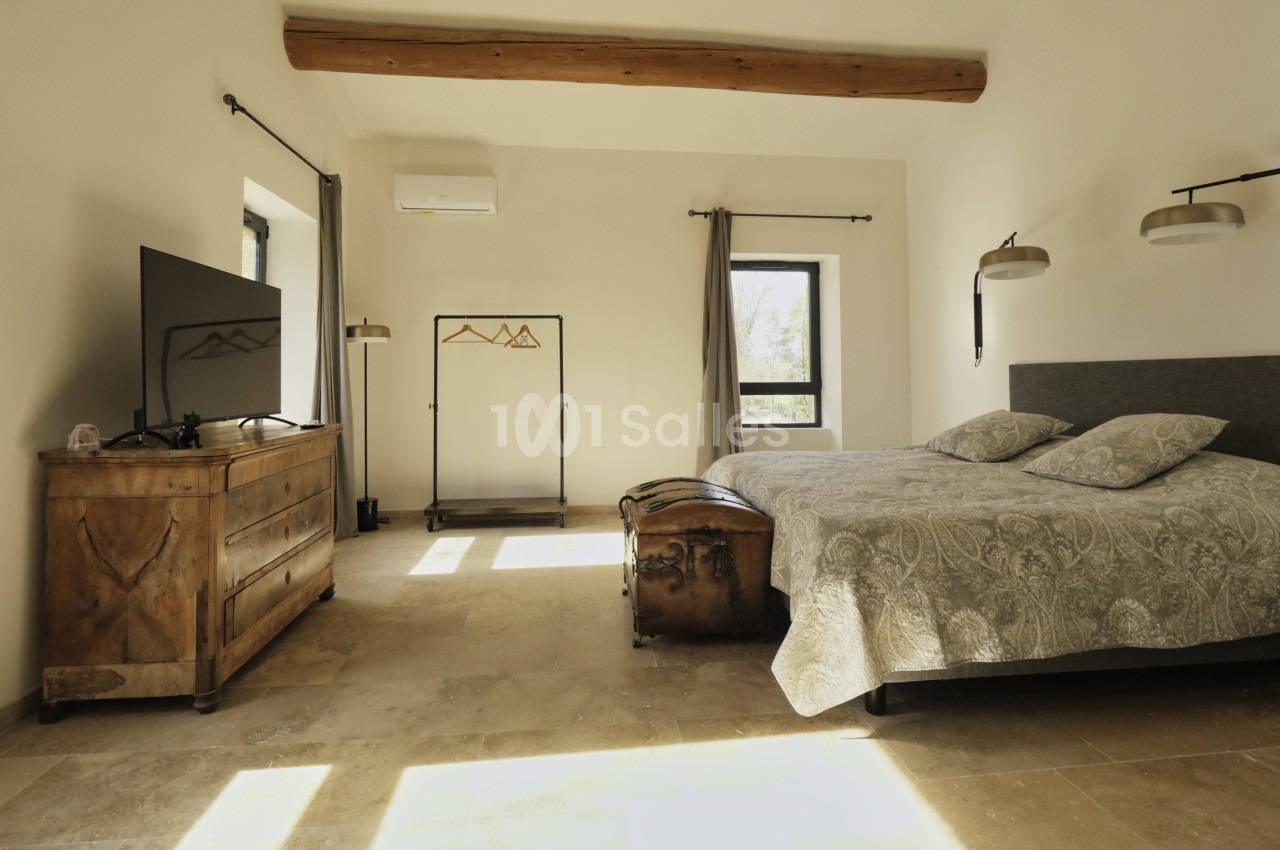 Chambre lumineuse avec lit double, commode en bois, portant à vêtements, télévision et poutre apparente au plafond.