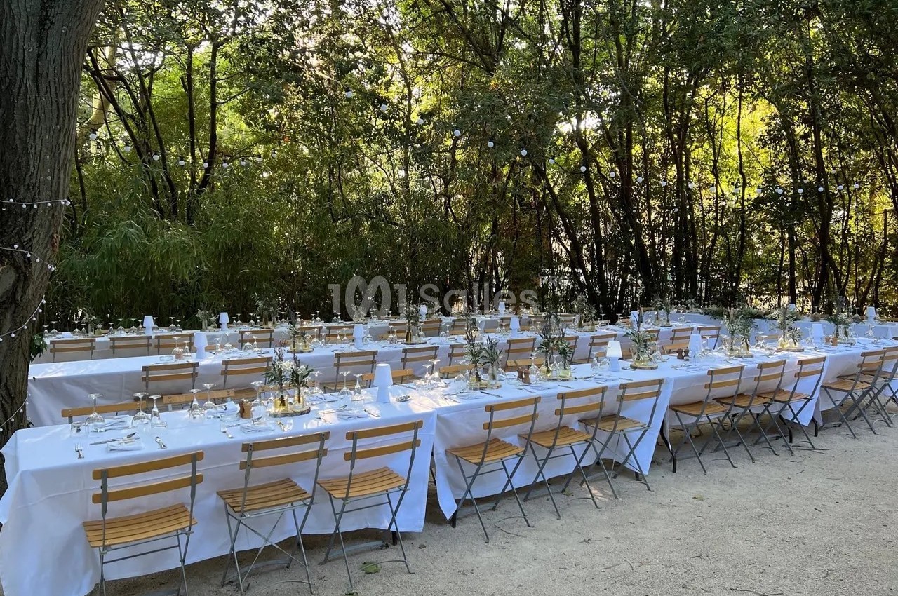 Tables longues dressées avec nappes blanches et chaises en bois, installées en extérieur sous des arbres.