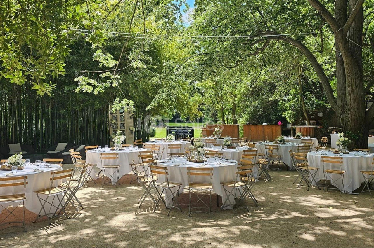 Tables rondes dressées avec nappes blanches et chaises en bois dans un jardin ombragé, entouré d'arbres et de verdure.
