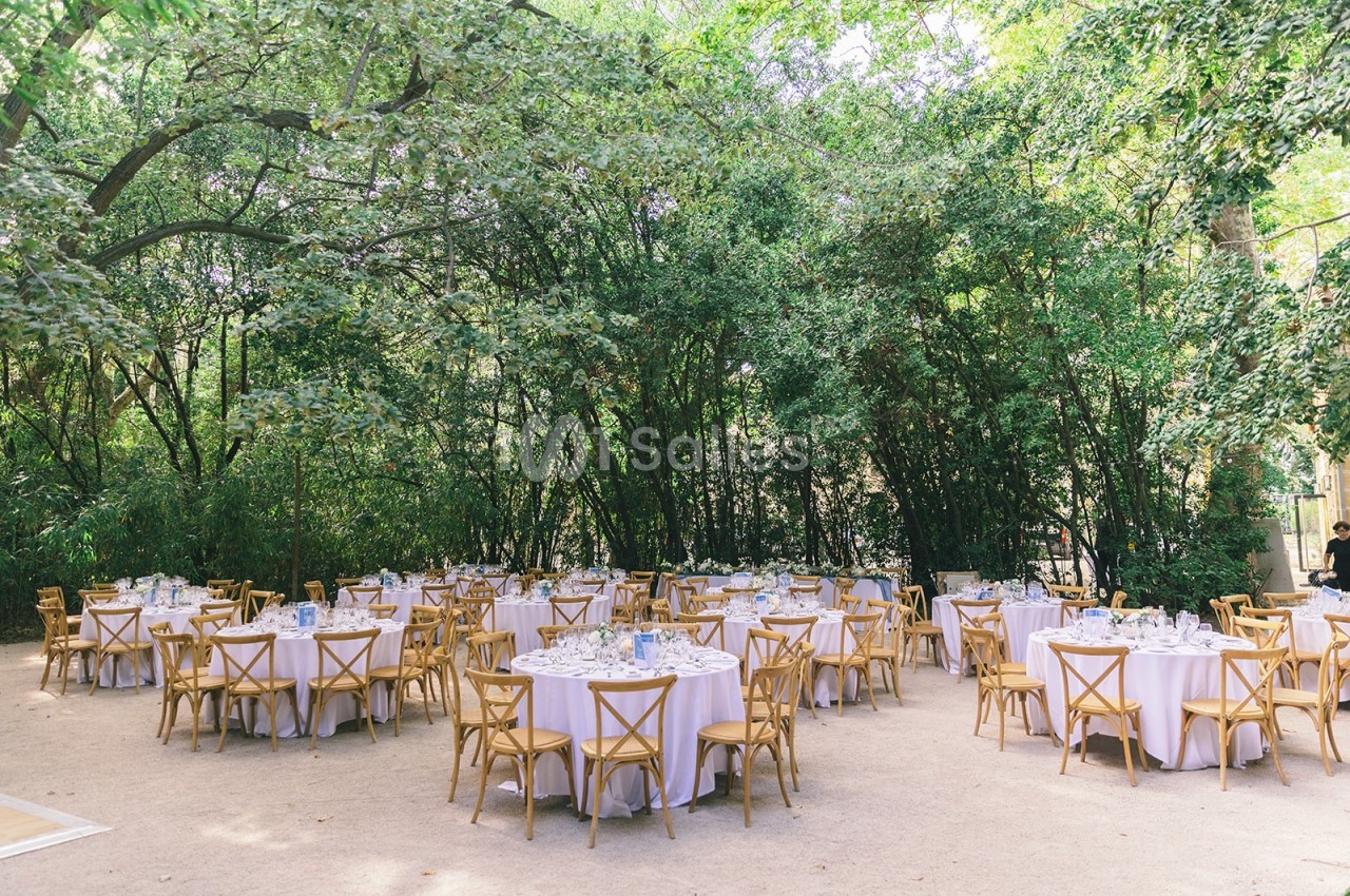 Tables rondes dressées avec nappes blanches et chaises en bois, disposées en extérieur sous des arbres verdoyants.