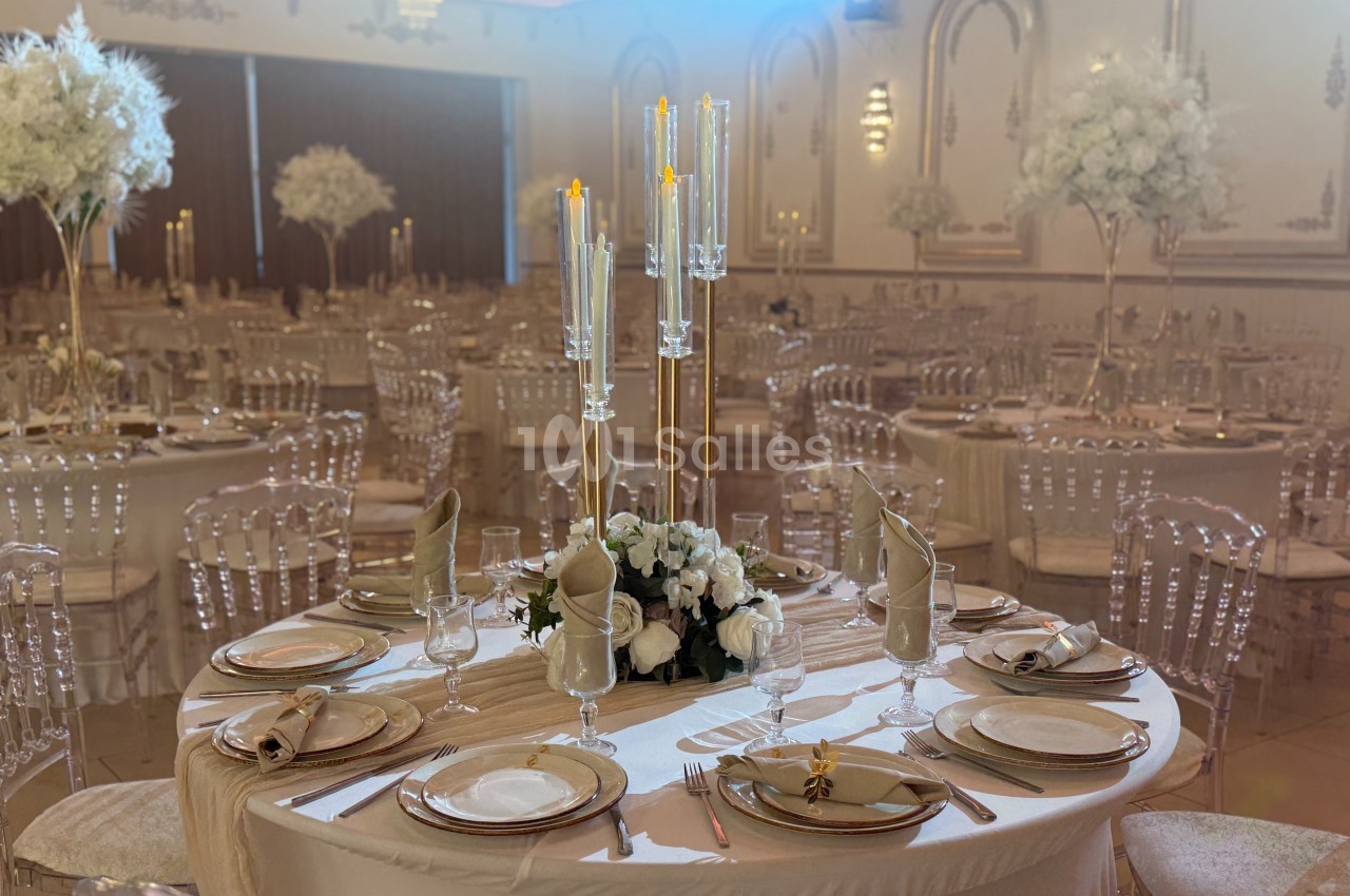 Salle de réception élégante avec tables rondes dressées, chandeliers, nappes blanches et décorations florales blanches.