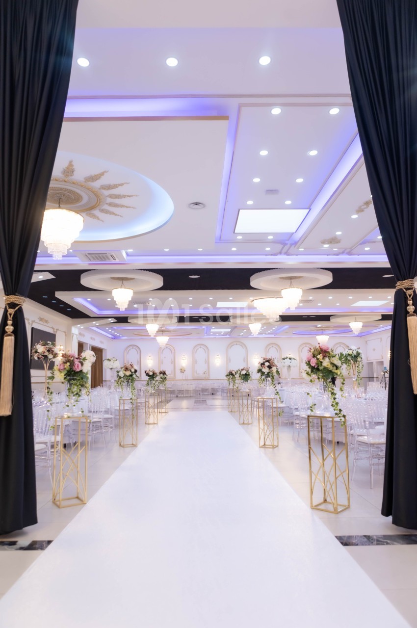 Salle de réception élégante avec allée centrale bordée de fleurs, éclairage lumineux et décorations raffinées.