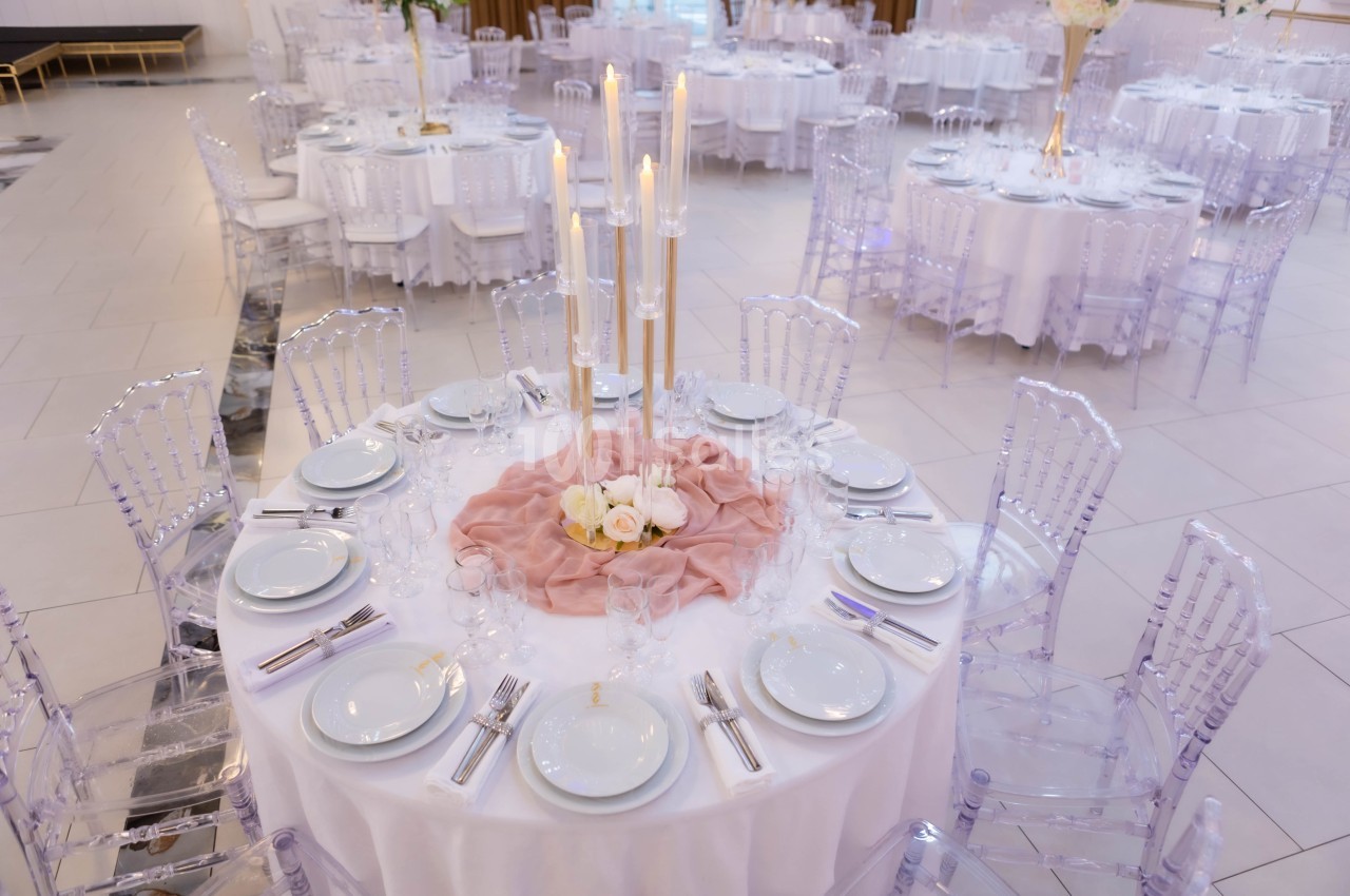 Table ronde élégamment dressée avec nappes blanches, chaises transparentes, centre de table floral et bougies hautes.