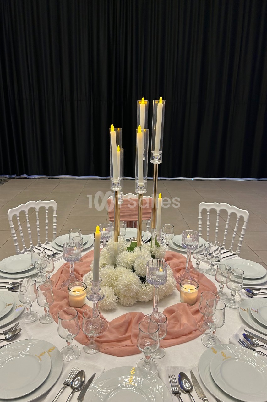 Table élégamment dressée avec nappes blanches, chemin de table rose, chandeliers hauts et décorations florales blanches.