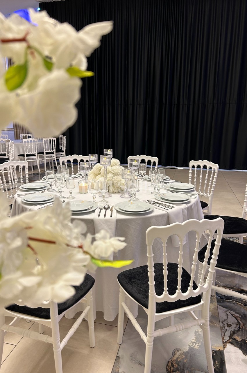 Table dressée avec nappes blanches, vaisselle élégante et centre de table floral, entourée de chaises blanches.