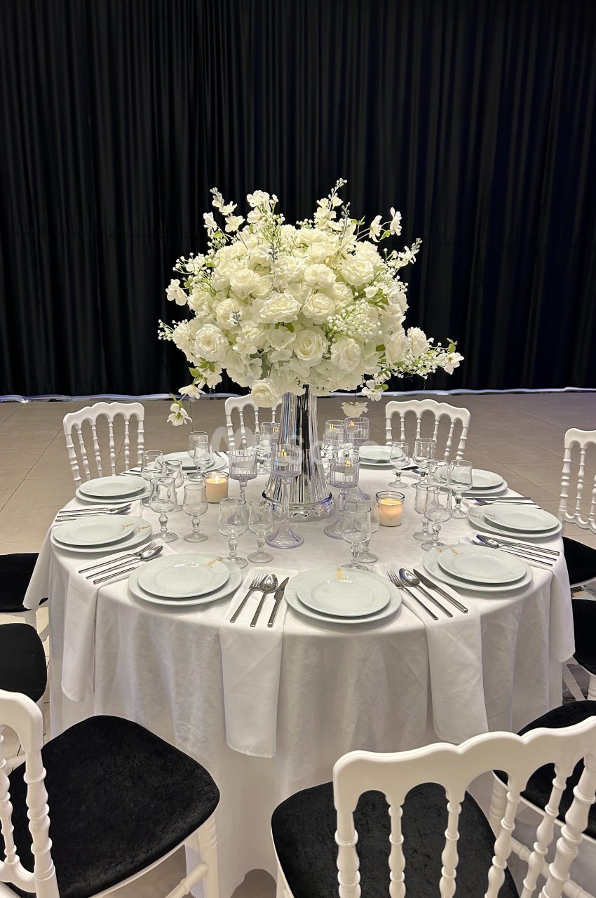 Table ronde élégamment dressée avec une nappe blanche, des couverts alignés et un grand bouquet de fleurs blanches au centre.