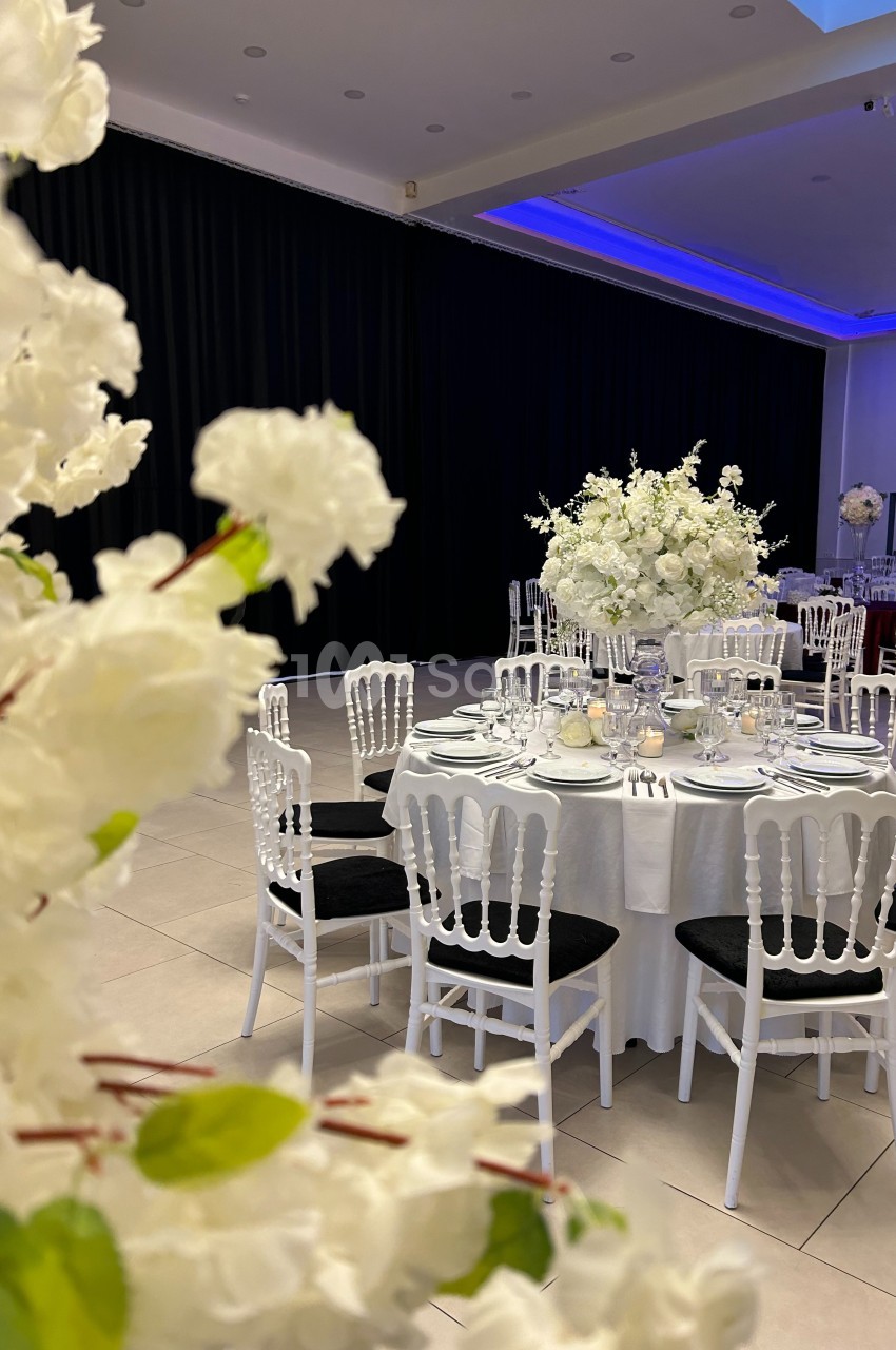 Salle de réception élégante avec tables rondes dressées, décorées de fleurs blanches et chaises blanches.