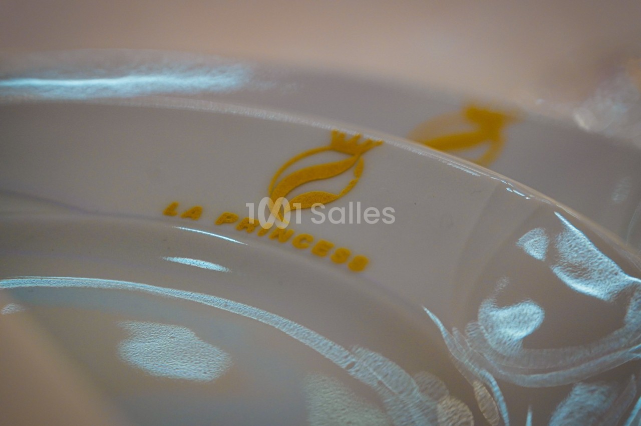 Assiette blanche avec un logo doré représentant une couronne et une flamme, accompagnée du texte ’La Princess’.