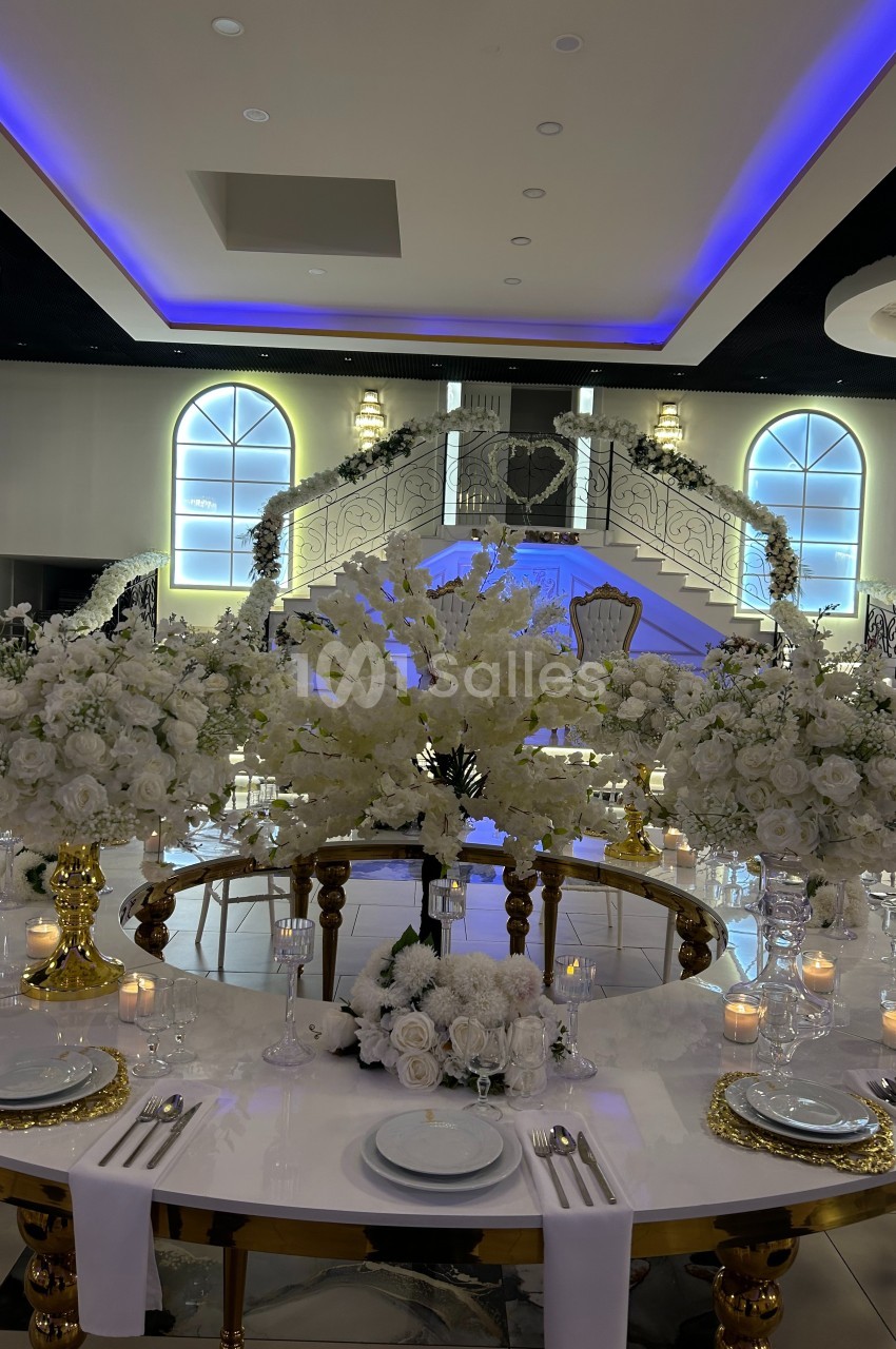Salle de réception élégante avec table décorée de fleurs blanches, chandelles et escalier illuminé en arrière-plan.