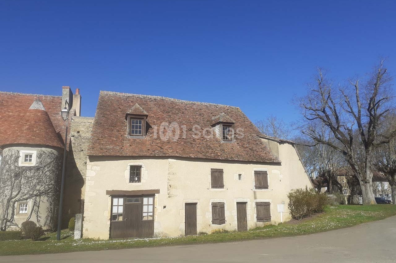 Maison ancienne en pierre avec toit en tuiles, entourée d'arbres et située au bord d'une route sous un ciel bleu.