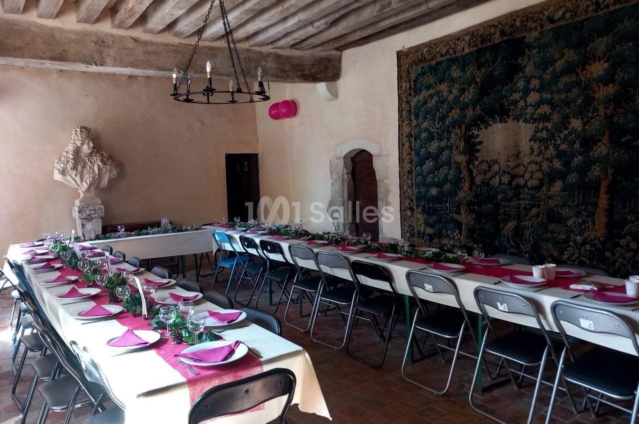 Salle rustique avec tables dressées pour un repas, nappes blanches, serviettes roses, tapisseries et lustre ancien.