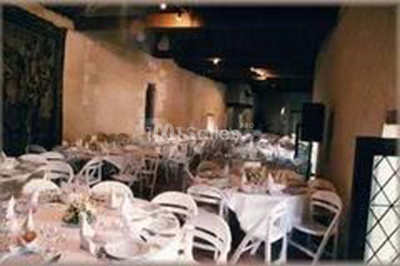 Salle de réception avec des tables rondes dressées et des chaises blanches, éclairée par une lumière tamisée.