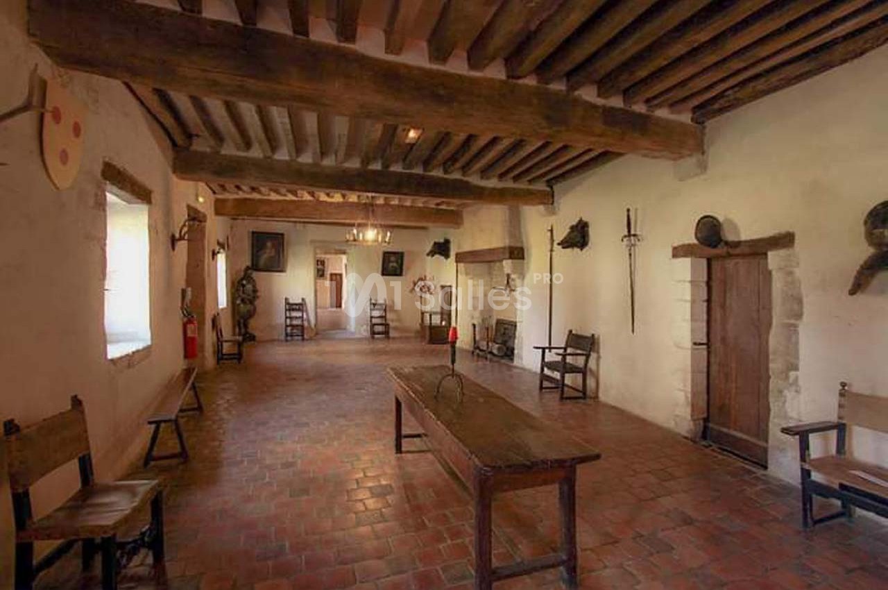 Salle ancienne avec poutres en bois, sol en terre cuite, mobilier rustique et décoration médiévale.