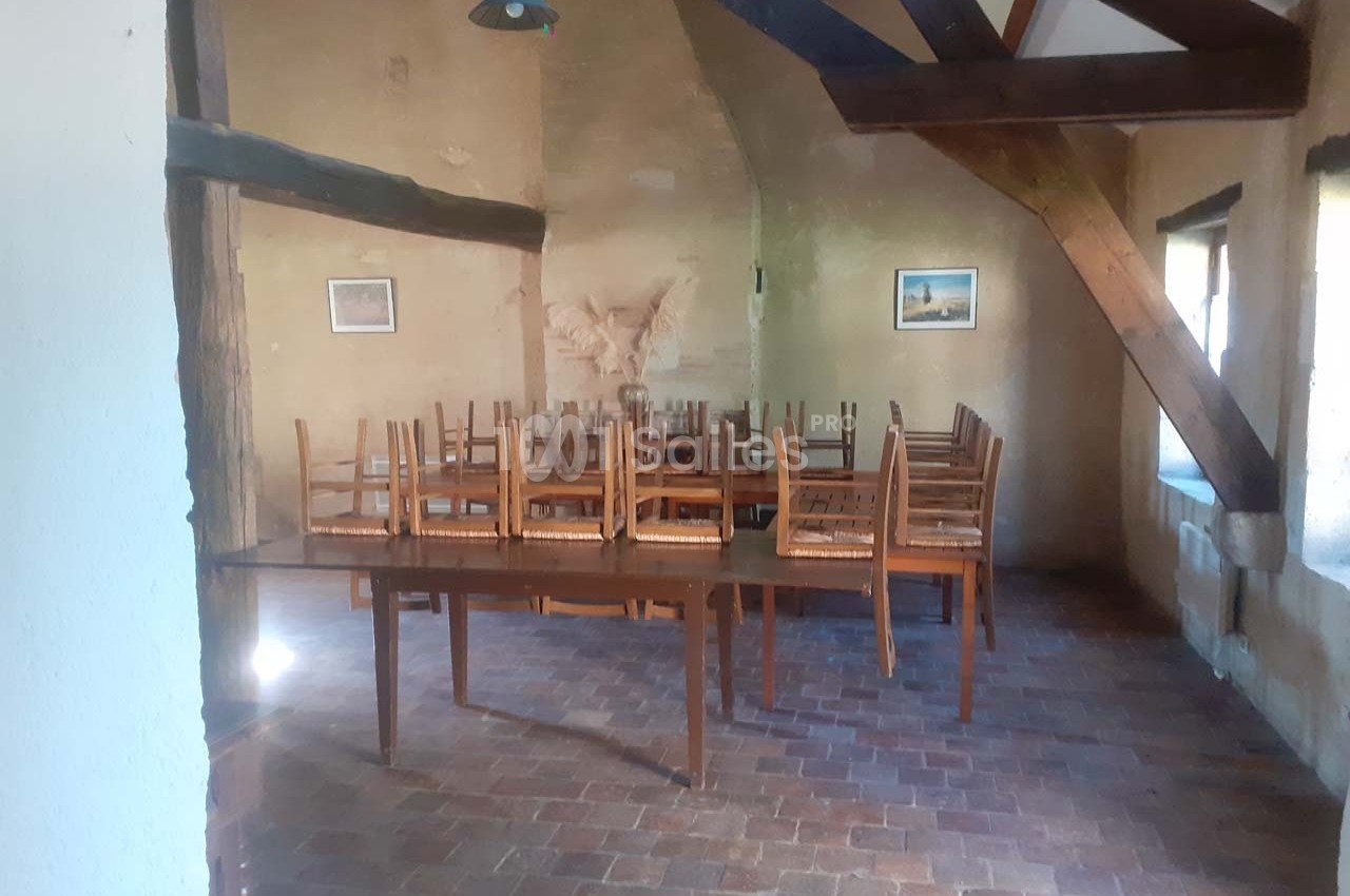 Salle rustique avec poutres apparentes, tables en bois et chaises empilées, éclairée par des fenêtres latérales.